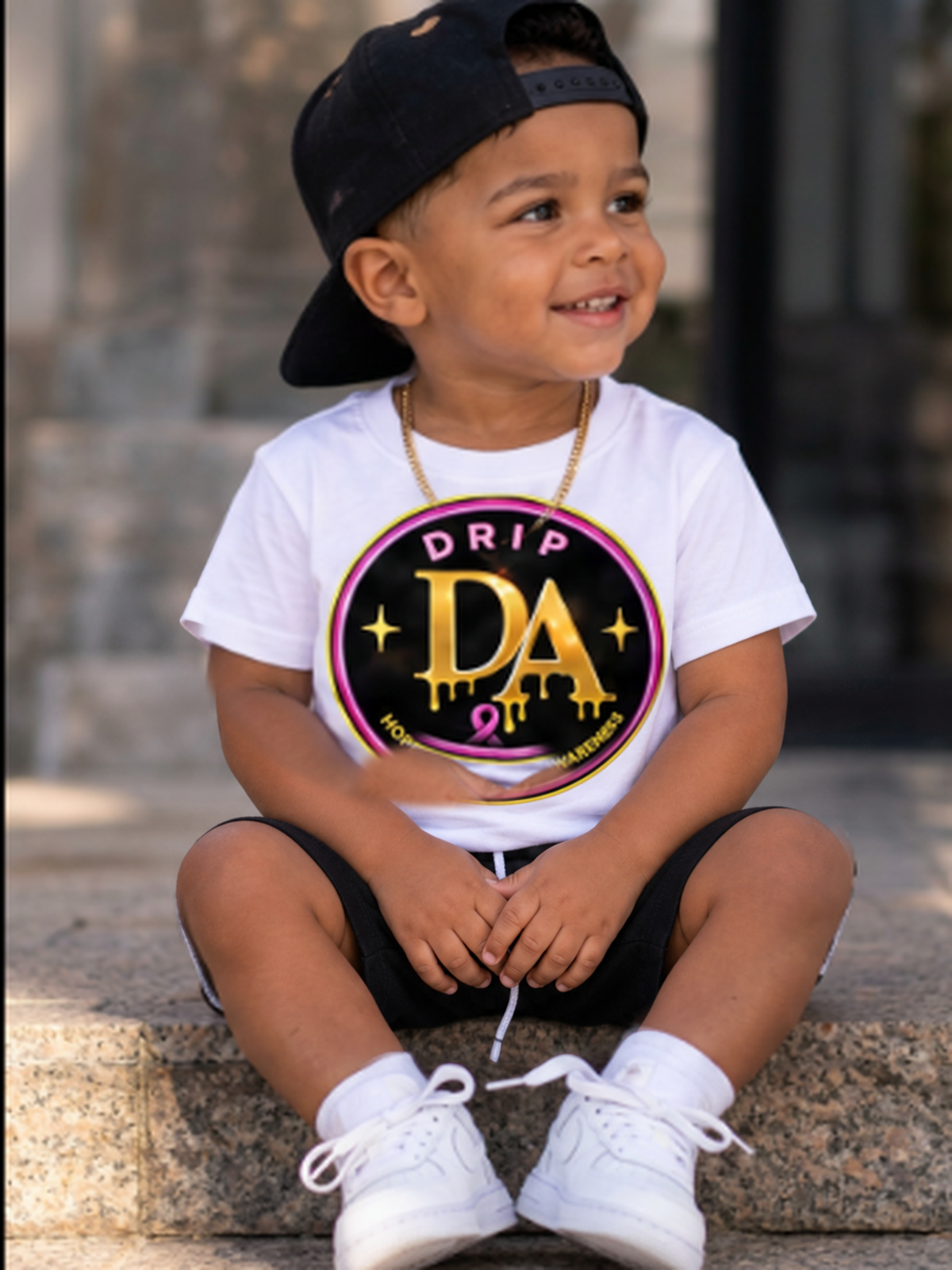 Toddler boy seated in a white Drip Avenue DA awareness shirt, black shorts, black cap, and white sneakers